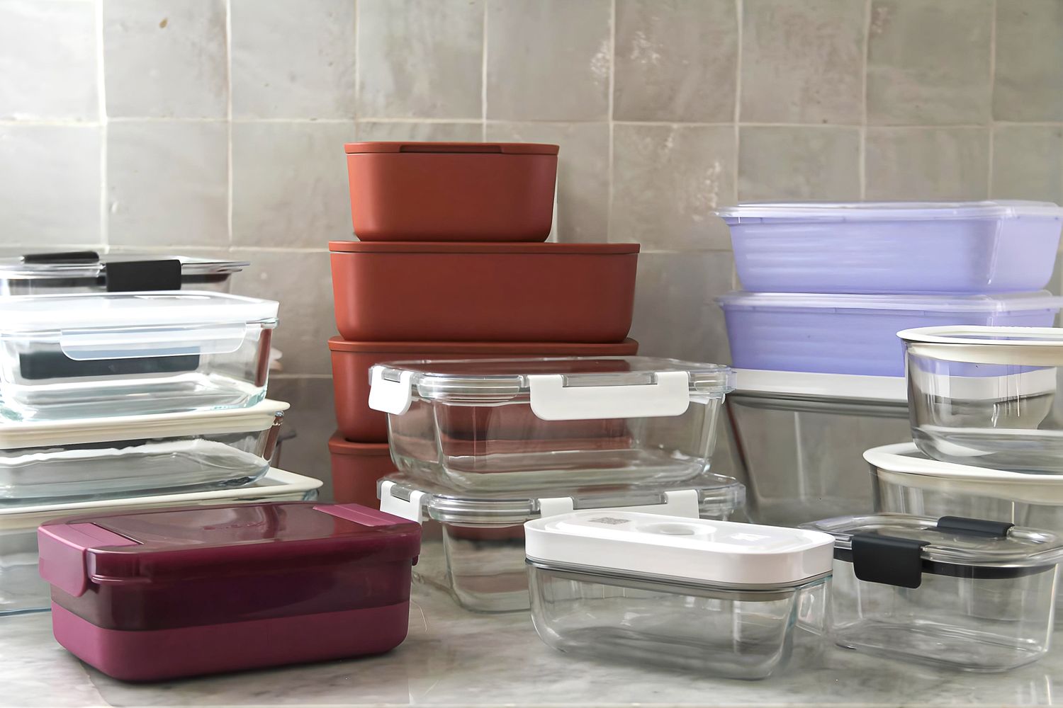 A collection of various food storage containers in different shapes and sizes stacked on a kitchen counter against a tiled backsplash