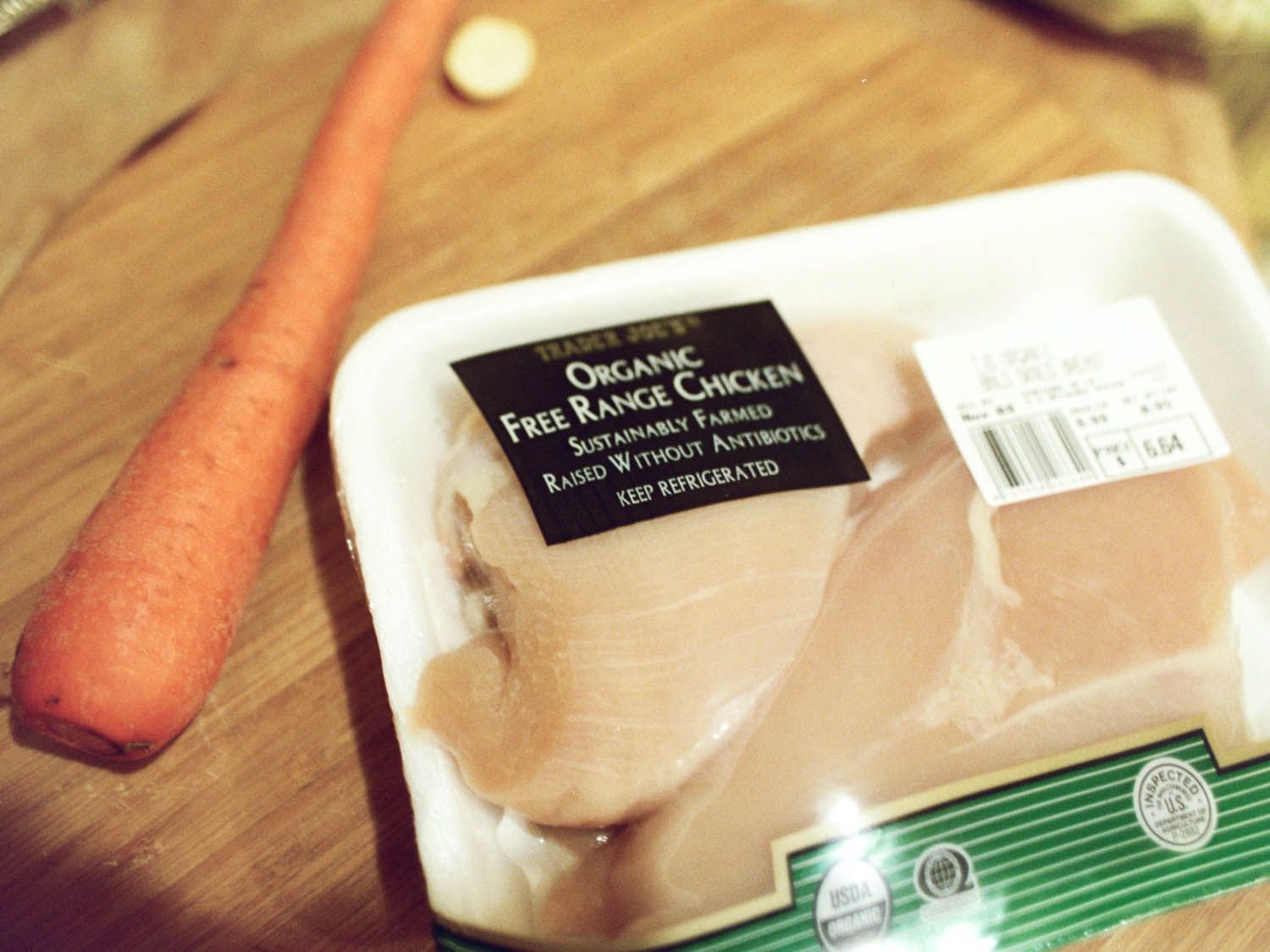 A package of organic skinless chicken breasts on a wood cutting board next to a carrot.