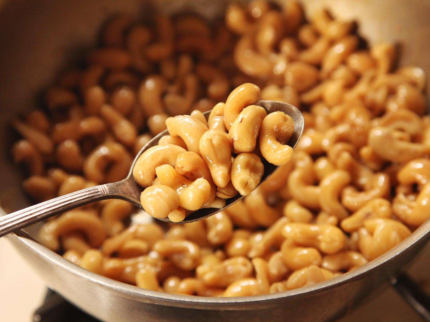 Spoonful of cooked cashews with a large pan of cooked cashews in the background.