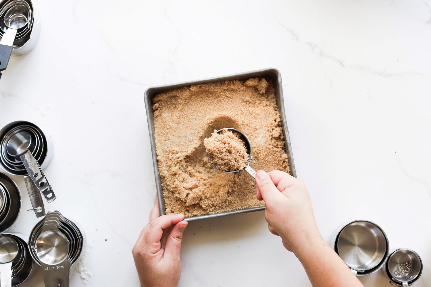 A person using a measuring cup to scoop brown sugar out of a baking dish.