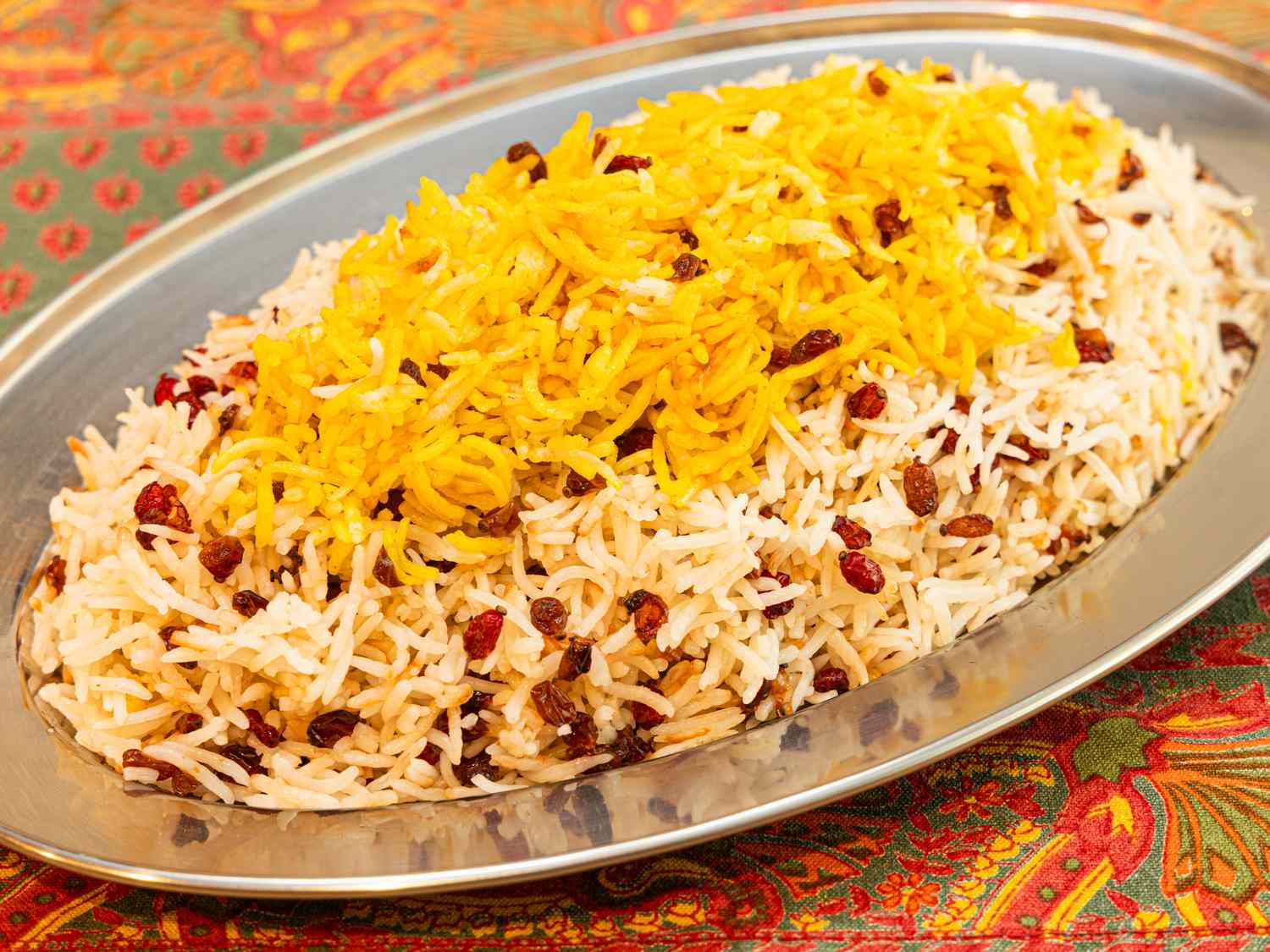 A dish of rice topped with barberries served on a metal platter placed on a patterned tablecloth