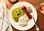 A plate with mashed peas, mashed potatoes, a sausage, and a fork resting on the edge