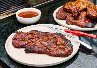 Cooked pork steak on a plate with barbecue sauce and utensils additional steaks in the background on a platter on a black countertop