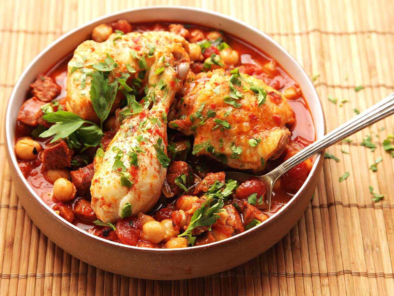 A serving bowl of Chicken With Chickpeas, Tomatoes, and Chorizo with a spoon.