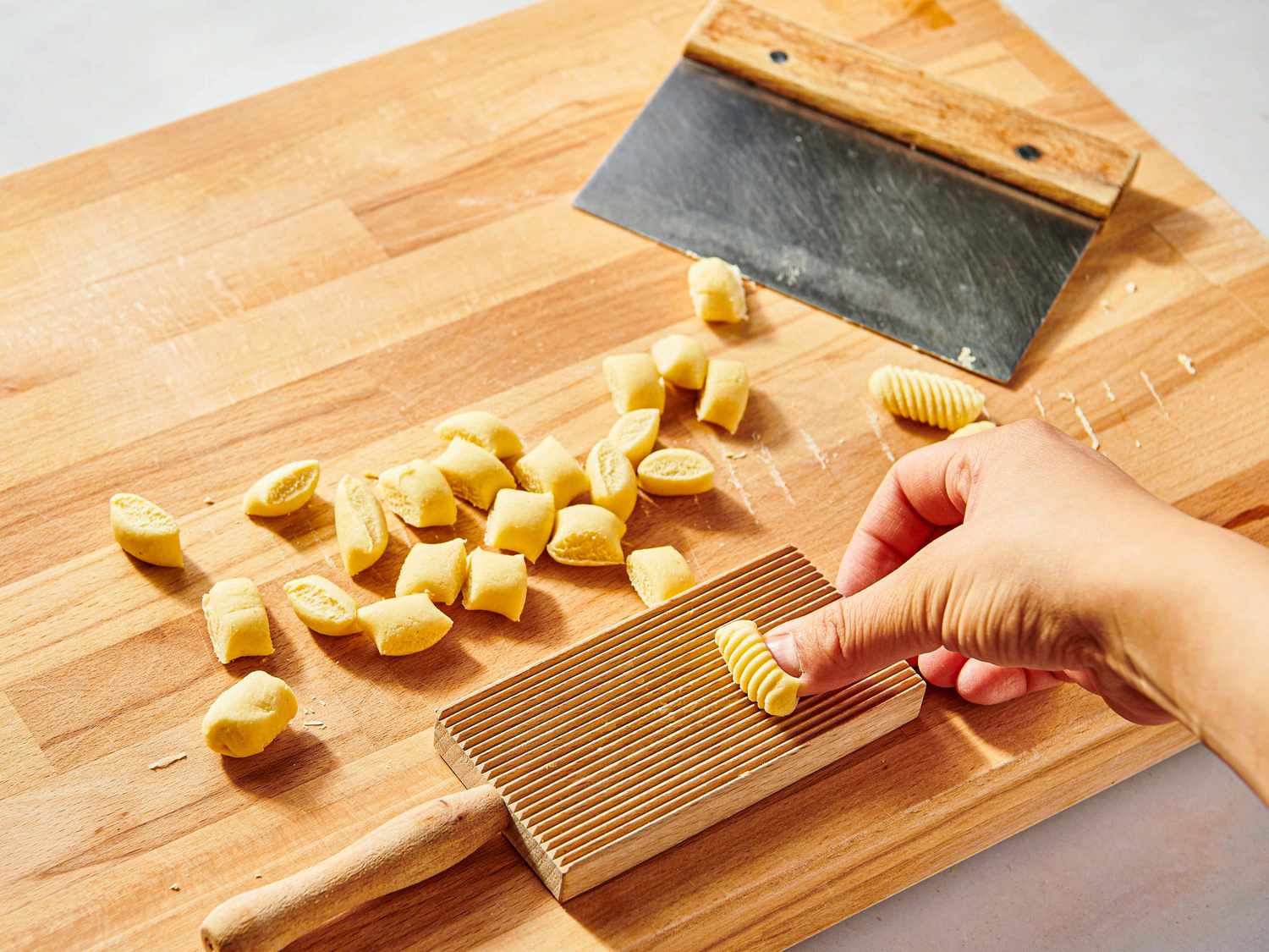 Hand pressing dough into gnocchi board on a wooden surface with other cut dough on it and a bench scraper 