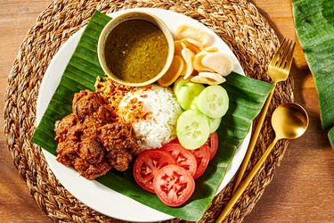 Plate of beef Rendang with rice, cucumbers, tomatoes and crisps on a banana leaf, with a woven placemat and gold cutlery