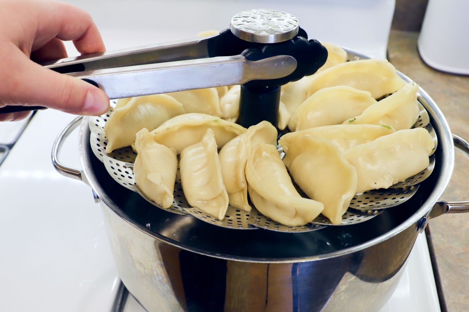 A person lifting a stainless steel steamer basket cooking dumplings out of a stockpot.