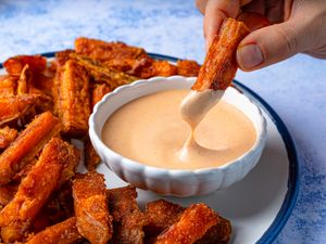 Hand dipping a sweet potato fry into a bowl of sauce sweet potato fries arranged on a plate