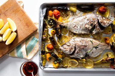 Overhead shot of 2 whole porgy roasted on a sheet pan with tomatoes, lemon slices, and shishito peppers