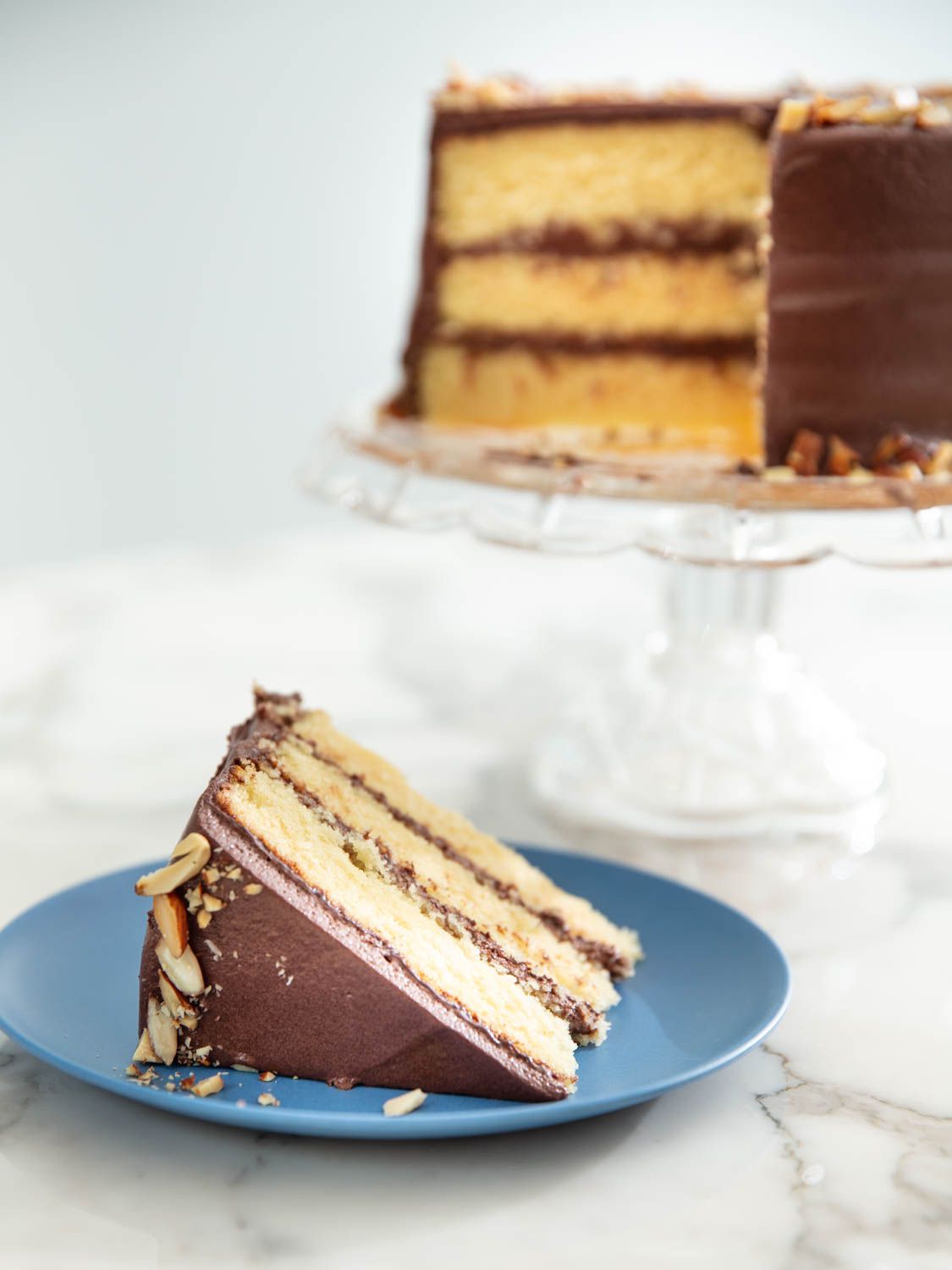 A slice of almond layer cake frosted with chocolate, with the remaining cake visible on a stand in the background