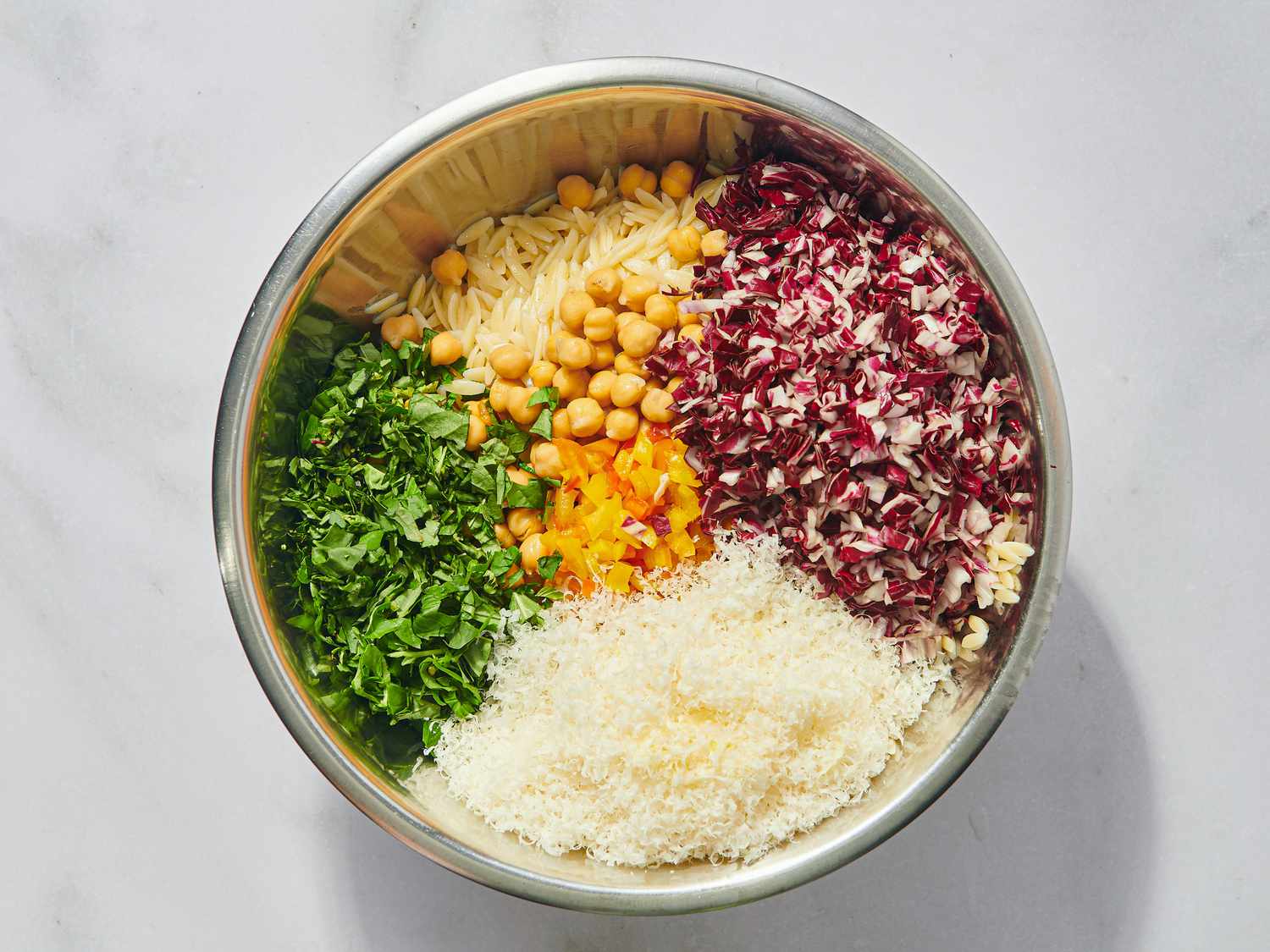 A metal bowl containing chopped greens, radicchio, chickpeas, grated cheese, and orzo pasta