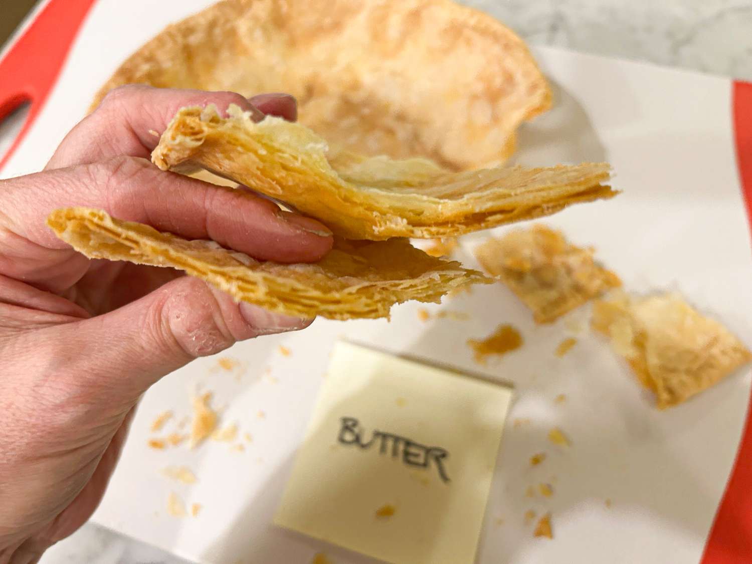 Hand holding a piece of layered pie crust butter and a cutting board in the background