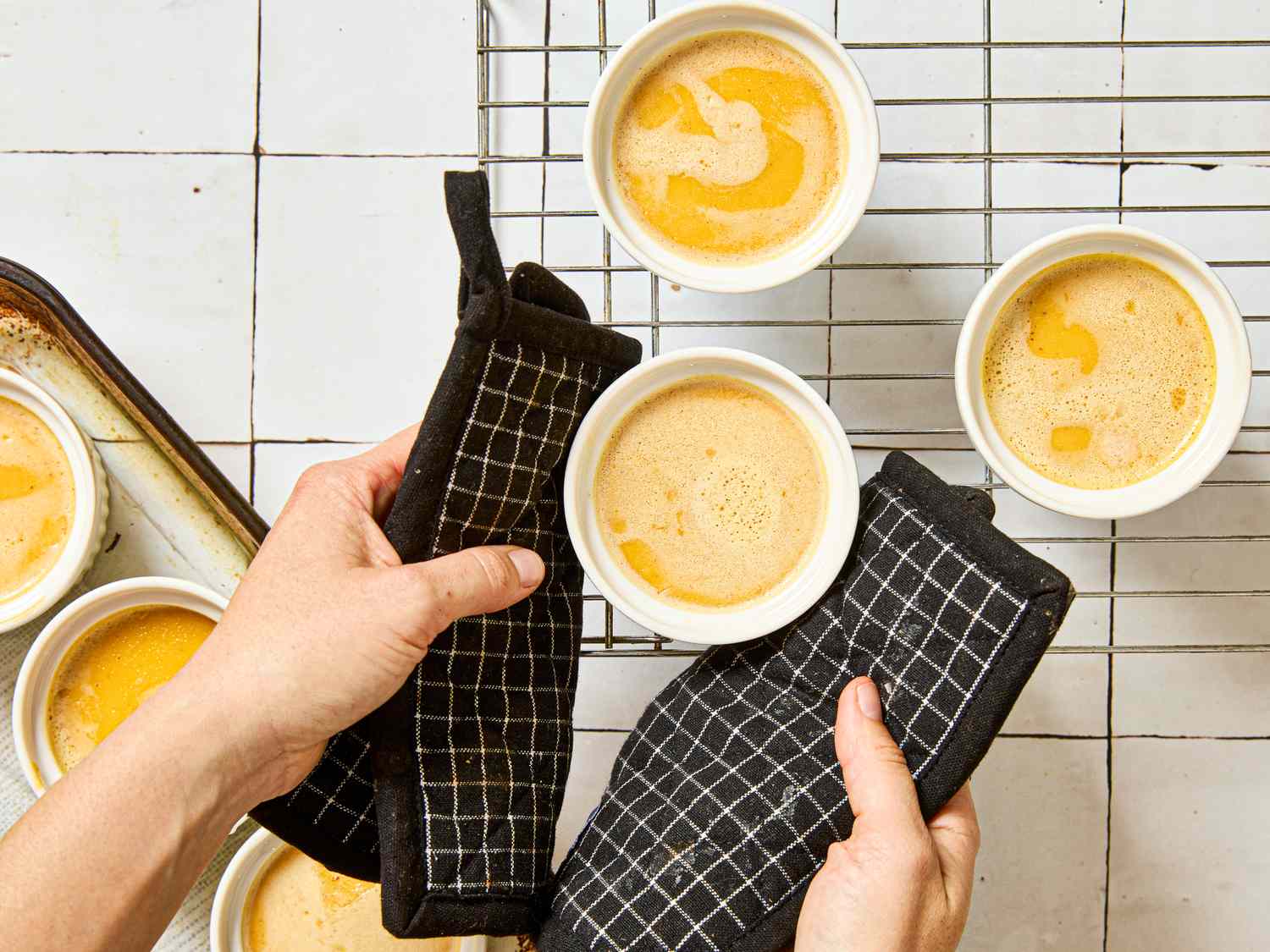 Hands in oven mitts holding ramekins of creme brulee on a cooling rack