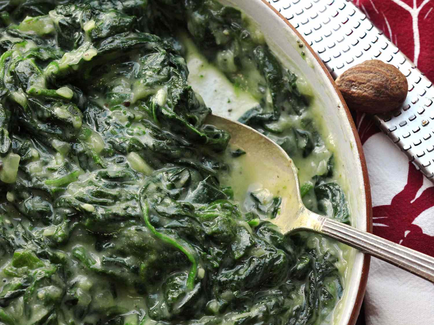 Vegan cauliflower creamed spinach in a casserole dish, with spoon, and a whole nutmeg and microplane next to it.
