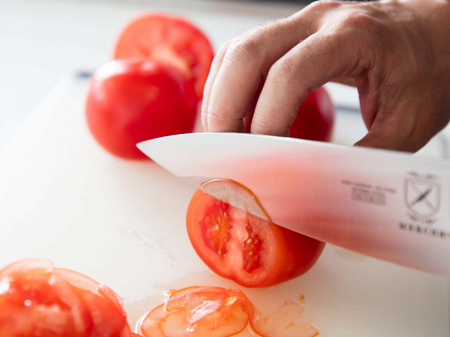 A small tomato is thinly sliced on a cutting board with a chef's knife.