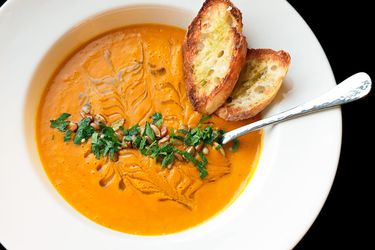 An overhead view of carrot soup, garnished with toasts, chopped fresh herbs, and a drizzle of olive oil. In a white bowl with a spoon.