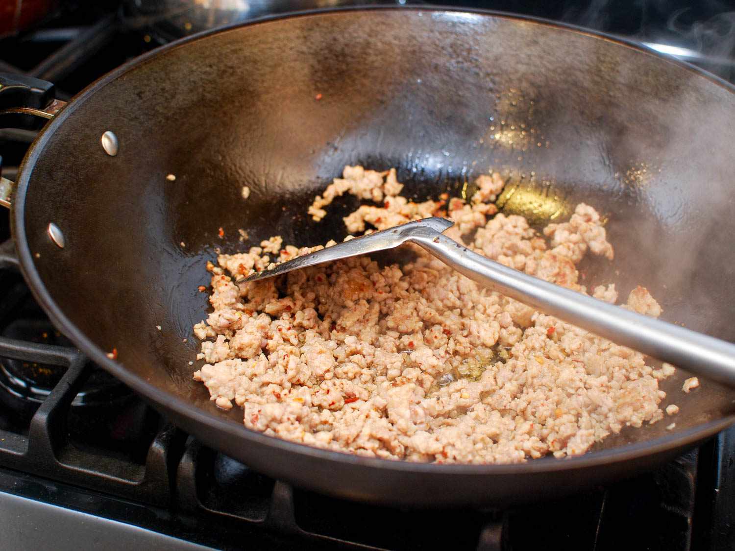 Ground pork being browned in a wok, tossed with a metal spatula. 