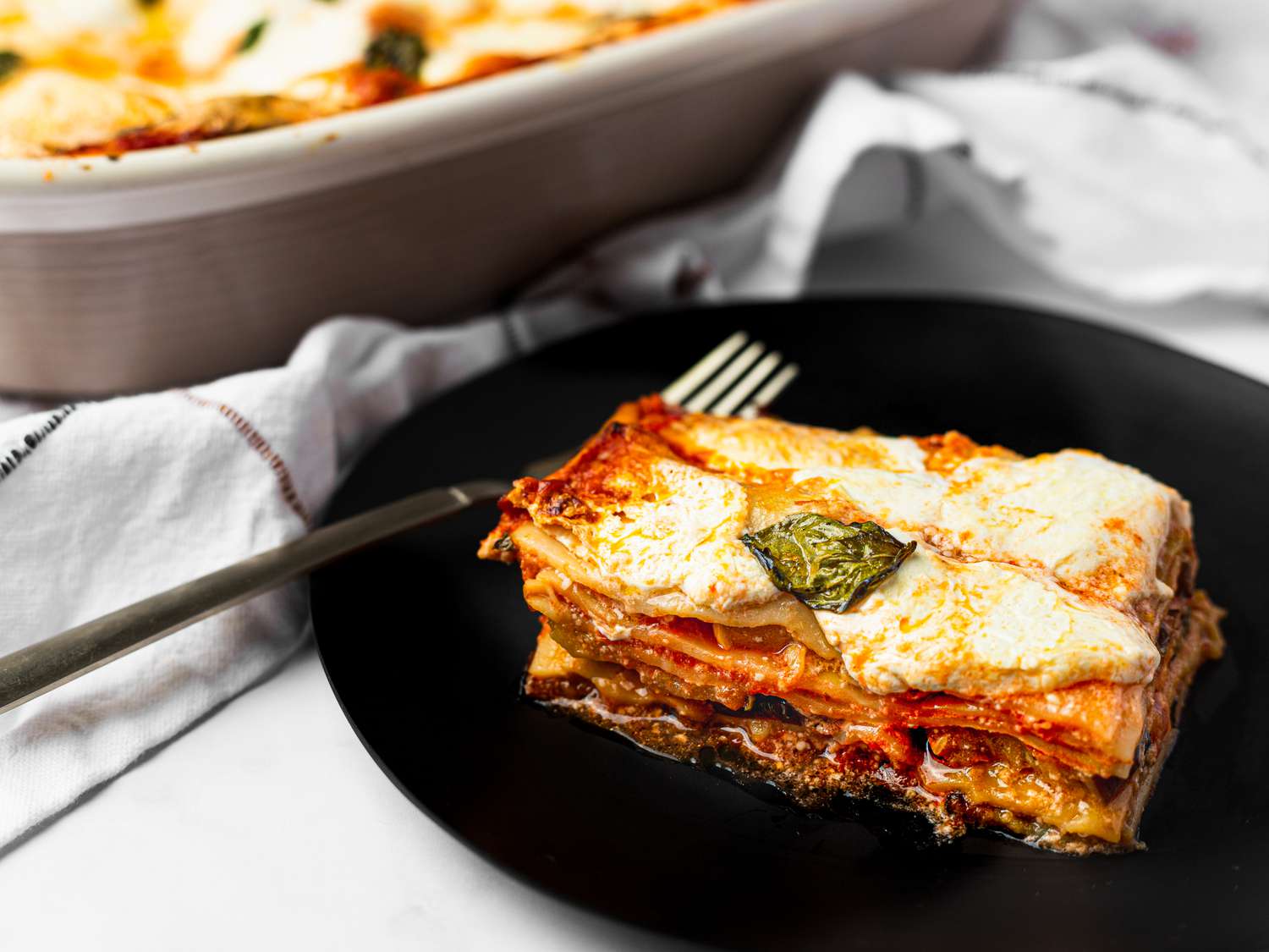 Slice of lasagna served on a small plate, with casserole dish holding lasagna peeking into the frame