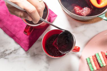 Ladle pouring mulled wine into red mugs, maroon napkin, and pink plate of christmas colored cookies