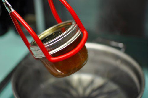 Red canning tongs lifting a small jar out of a pot.