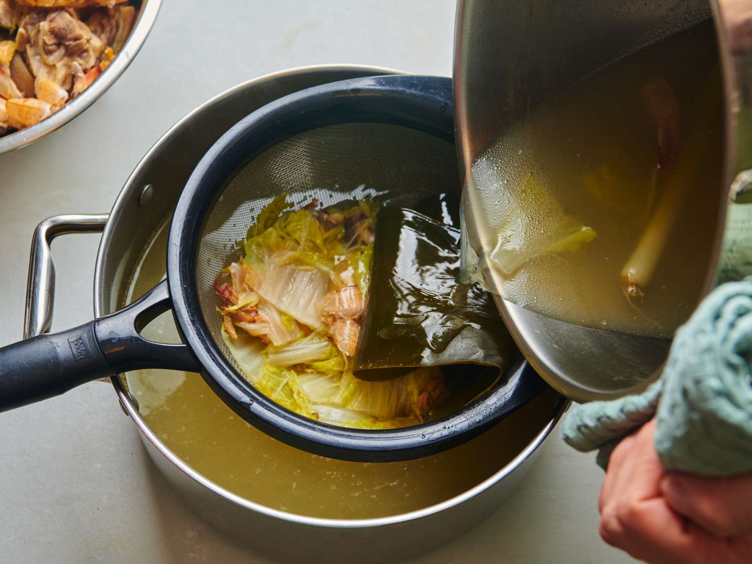 The broth being strained from the stockpot into another, smaller pot through a fine mesh strainer.