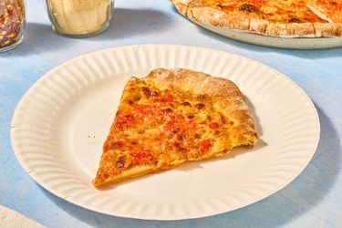 slice of mustard pizza on paper plate, and whole pizza in back, with parmesan and red pepper shakers 