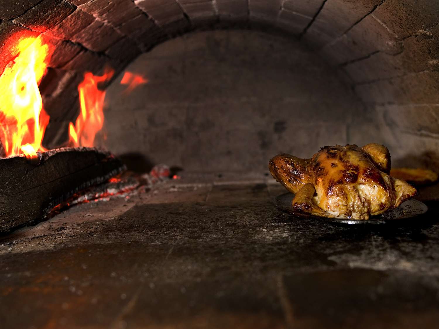 A roast chicken cooking in a brick woodfire oven with flames visible at the side