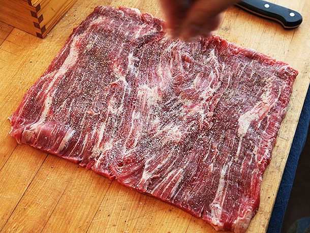 Large piece of butterflied flank steak being seasoned with salt and pepper