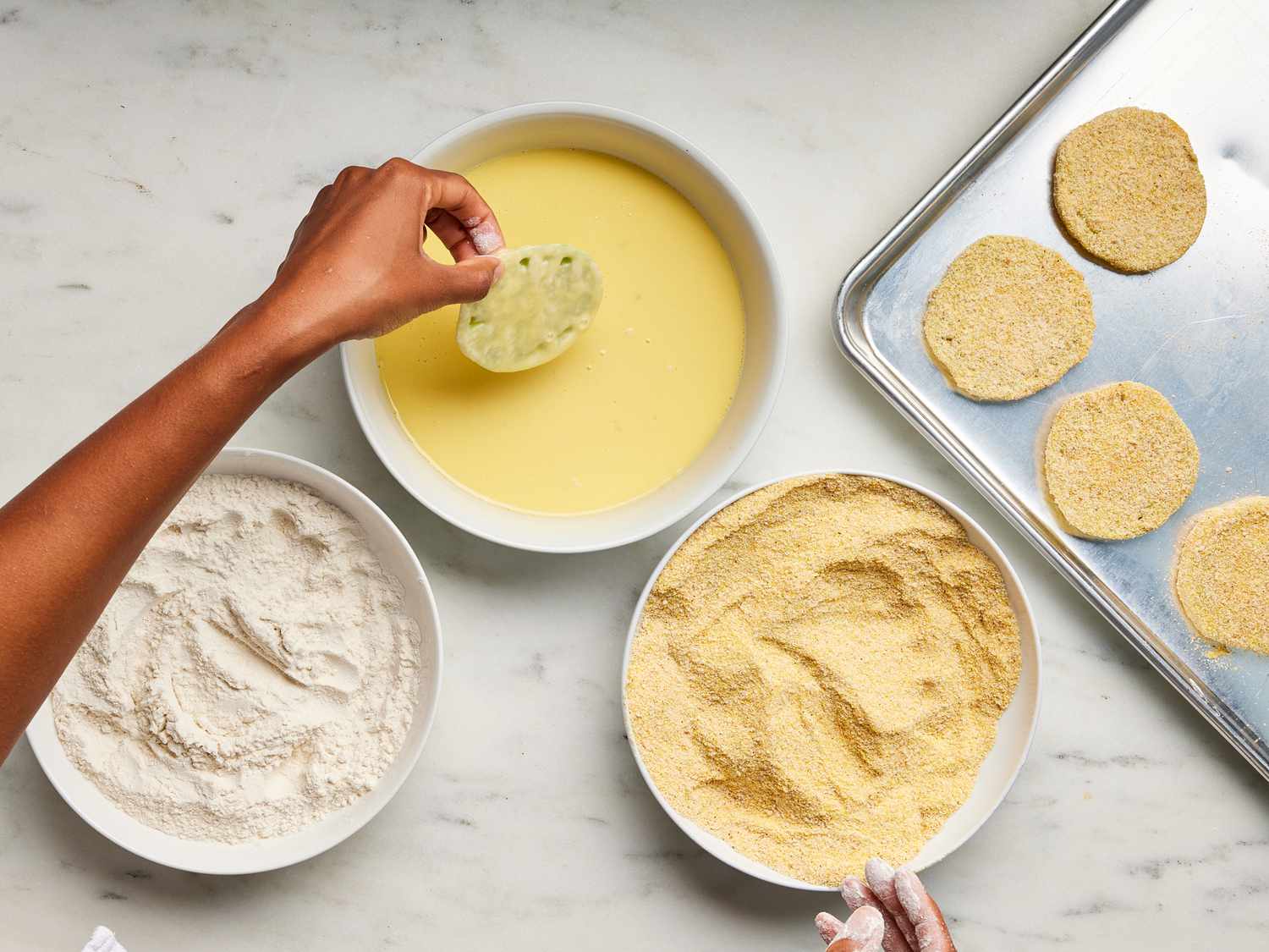 Cornmeal, egg, and flour in 3 separate bowls with sheet pan of dipped green tomatoes next to them. One tomato is being dipped in egg mixture