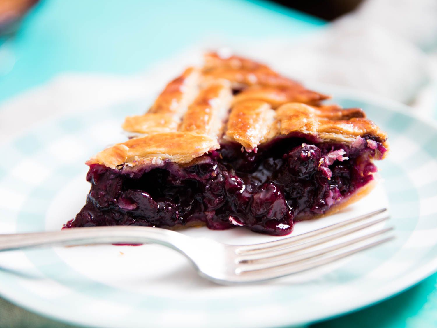 Close-up of a slice of blueberry pie on a serving plate, fork in foreground.