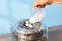 A person using a liquid measuring cup to add mini marshmallows through the top of the Cuisinart ice cream maker.