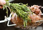 Boneless leg of lamb on a rotisserie, being basted with a bundle of fresh herbs. 