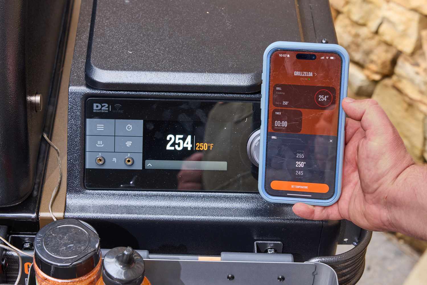 Closeup of a person holding a smartphone displaying a temperature control interface next to a grill with a digital temperature display
