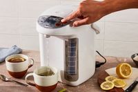 A person using a countertop water boiler to fill a cup with tea preparation items including lemon slices nearby