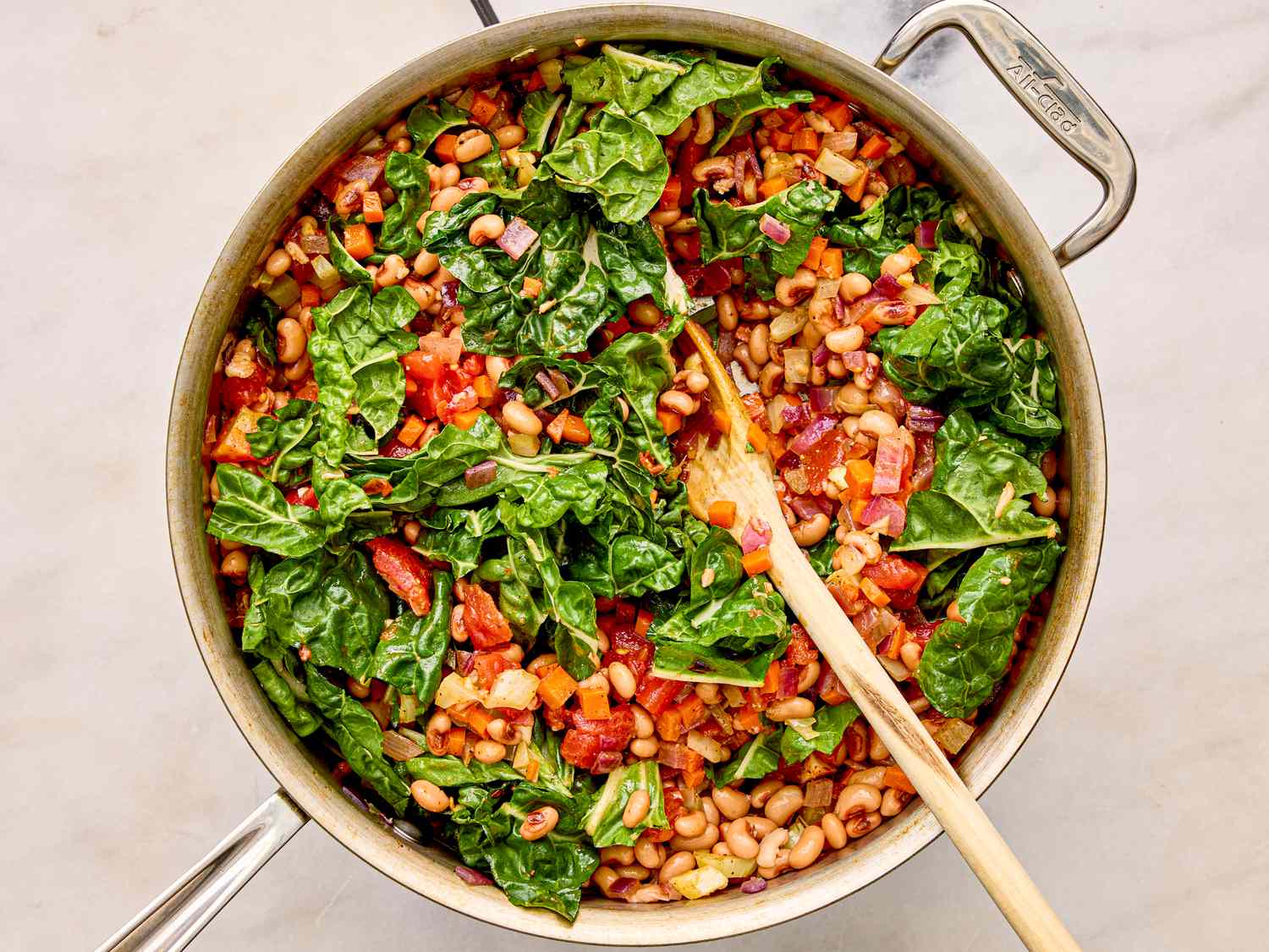 A pot containing a stew with blackeyed peas greens and other colorful vegetables being stirred with a wooden spoon
