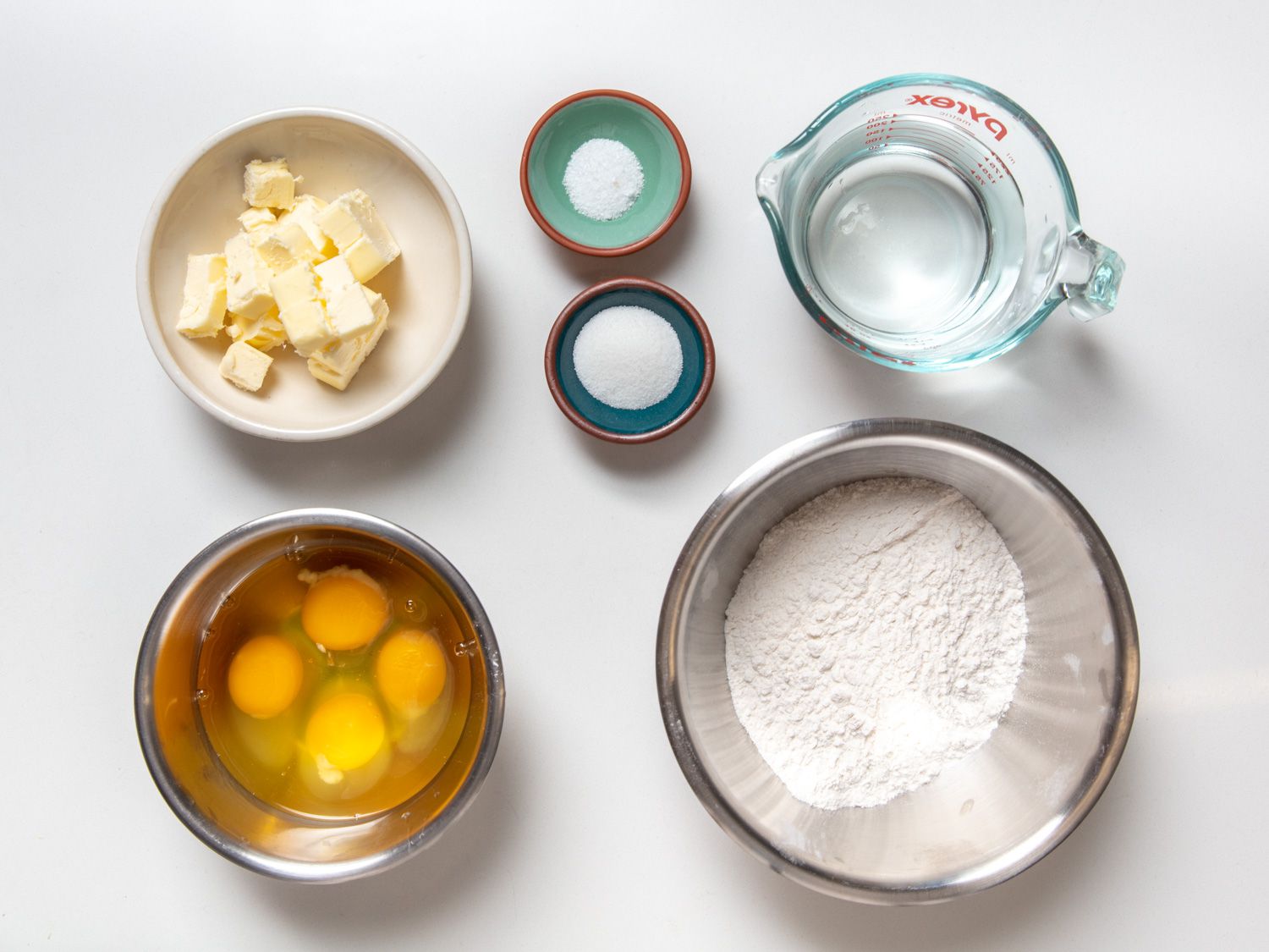 The key ingredients on choux pastry, in separate dishes on a counter: butter, water, eggs, flour, salt, and sugar