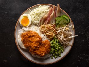 Overhead of a serving plate of noodles dressed with fish curry, with assorted vegetable garnishes and a soft-cooked egg on the side.