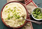 A bowl of creamy mashed potatoes in a red-brown bowl with sliced scallions on top