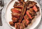 A t-bone (porterhouse) steak on a white plate, with the sliced meat placed around the bone. On the lefthand side of the image are several metal forks stacked on top of each other.