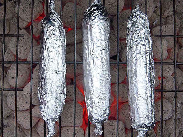 Overhead view of foil-wrapped ears of corn cooking over coals on a grill grate.