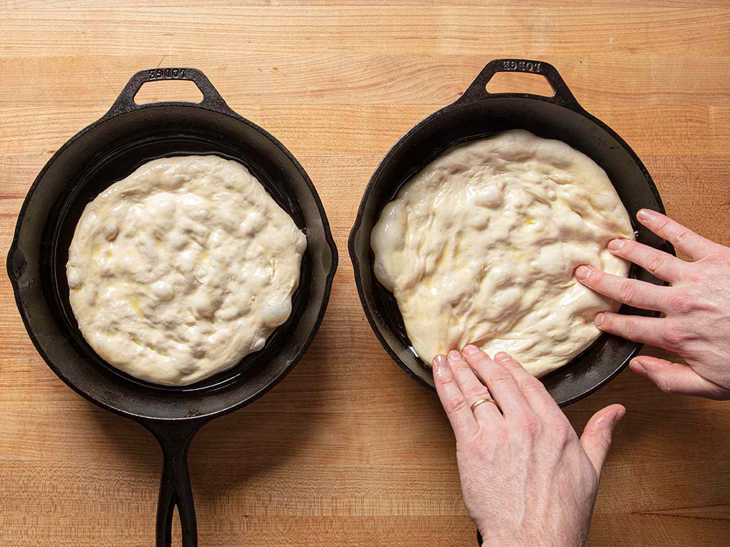 Two cast iron pans side by side containing pizza dough, with the rightmost pan having the dough spread out by a set of hands.