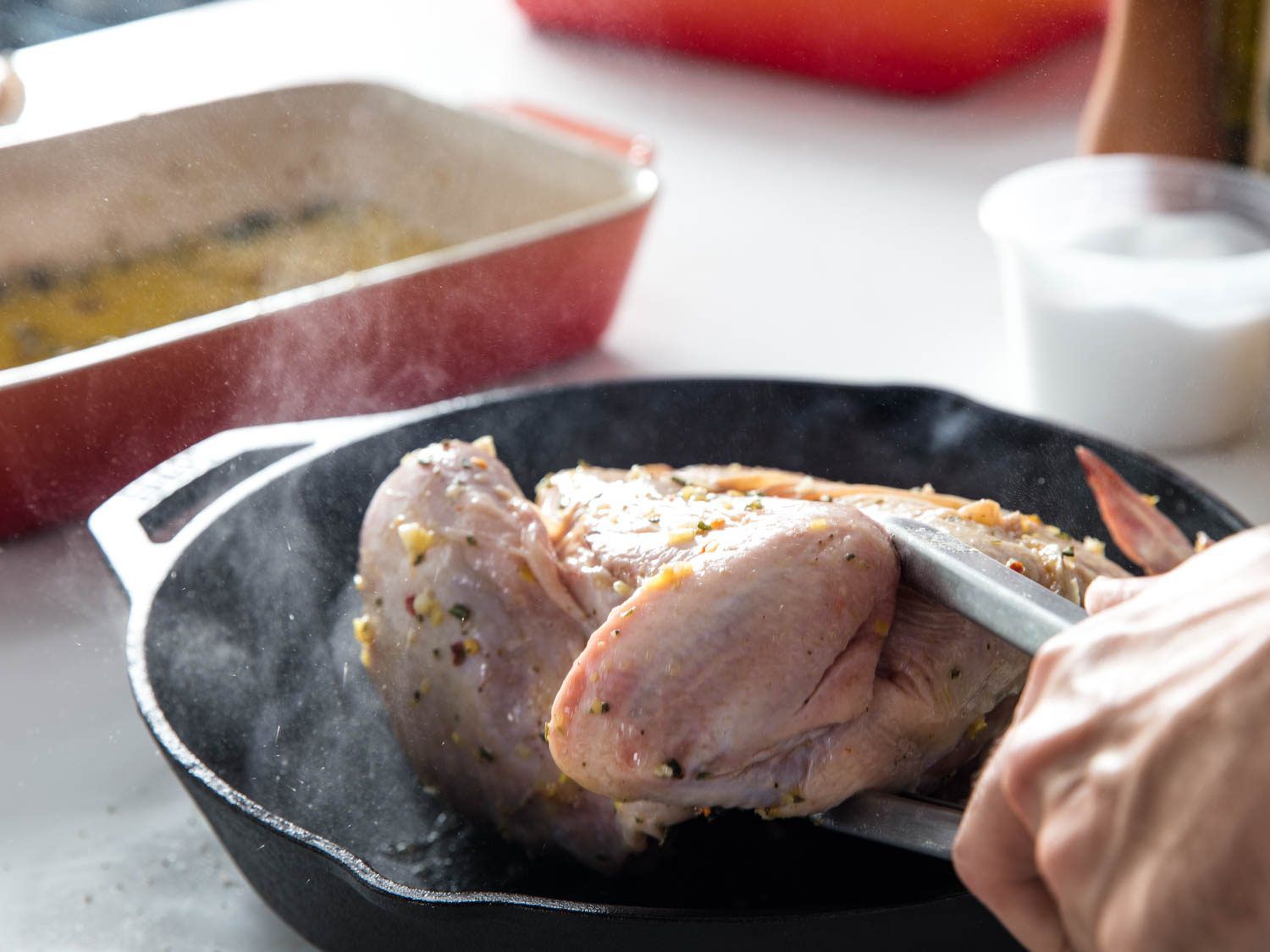 A hand using tongs to move a whole chicken from a dish with marinade to a cast iron skillet.