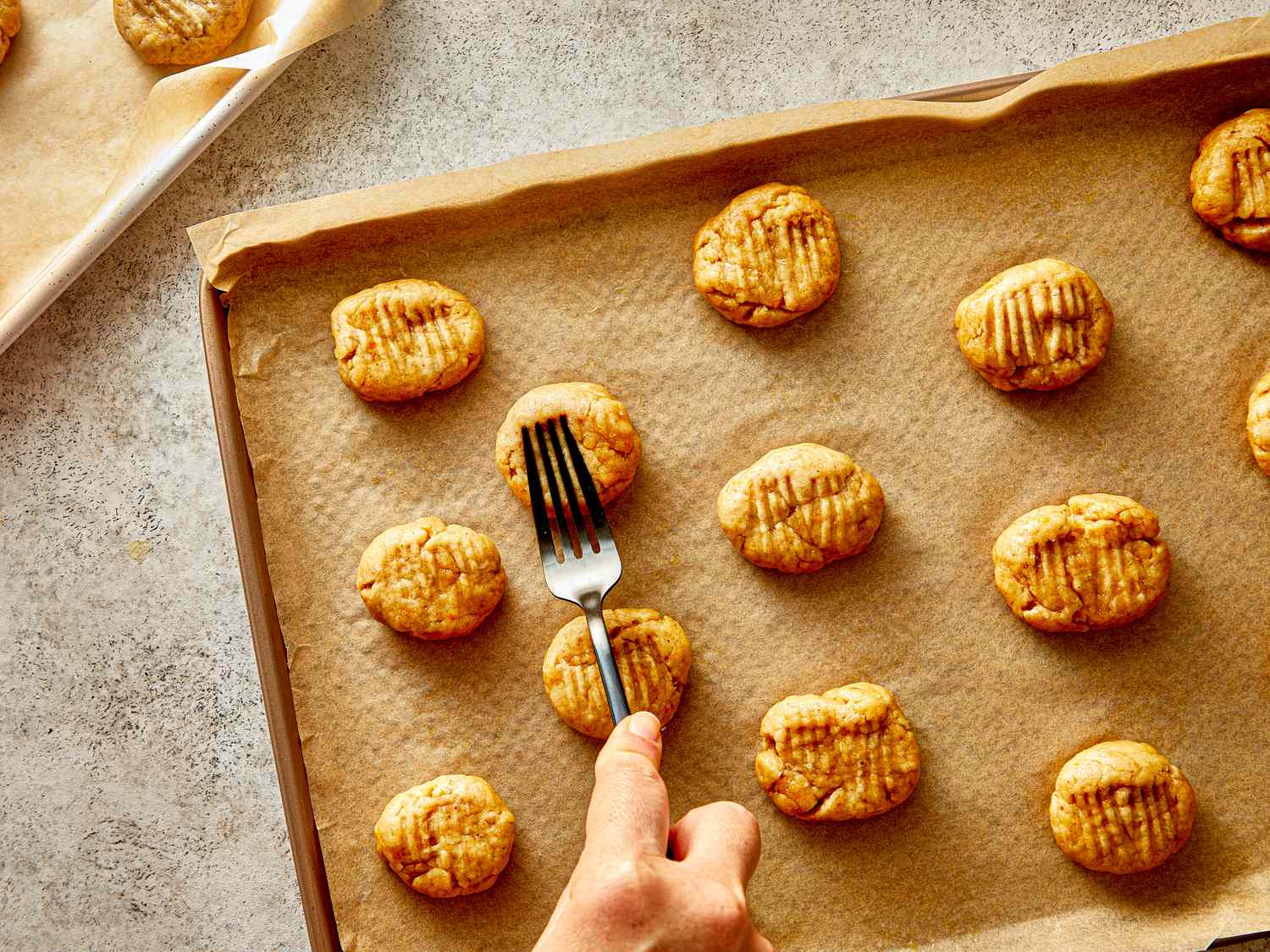 A hand pressing grooves on cookie dough balls with a fork on a baking tray lined with parchment paper