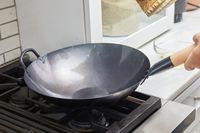 The Mammafong Pre-Seasoned Blue Carbon Steel Flat-Bottom Pow Wok displayed on a stove with a hand holding the handle