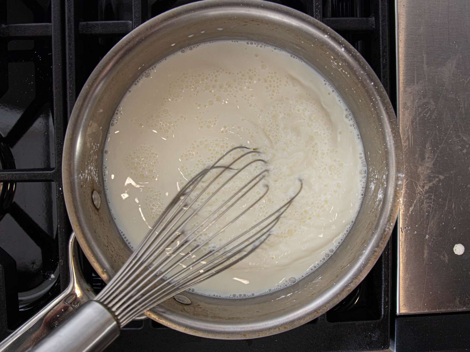 Whisk stirring a white mixture in a metal saucepan on a stove