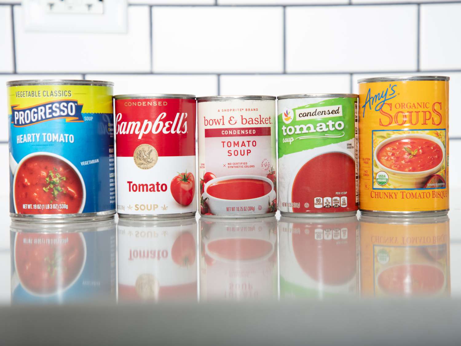 Five cans of tomato soup lined up on a surface each from a different brand labels visible