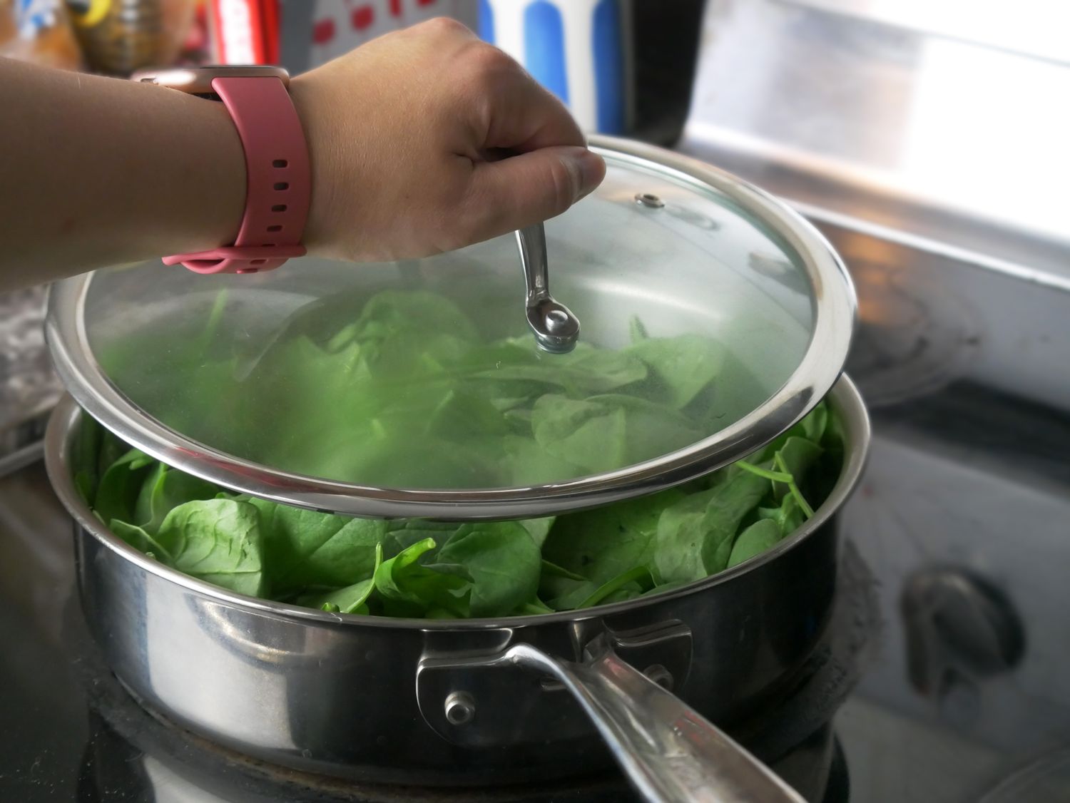 a person putting a glass lid on a saute pan filled with spinach