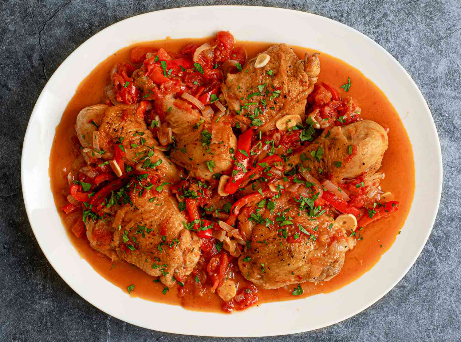  A white oval platter holding chicken cacciatore and a good amount of golden brown sauce. The platter is on a textured grey-blue surface.