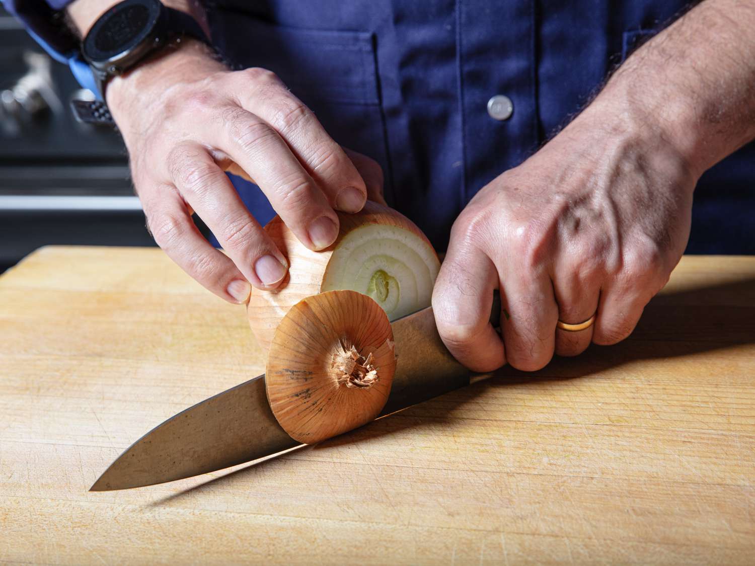 Hands slicing off the stem end of an onion on a cutting board.