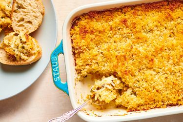 A blue baking dish holding golden brown Maryland Crab Imperial. The bottom left corner of the baking dish shows a metal spoon holding a spoonful of the crab, while on the lefthand side of the image is a light blue ceramic plate holding small pieces of bread, one of which has been topped with the crab.